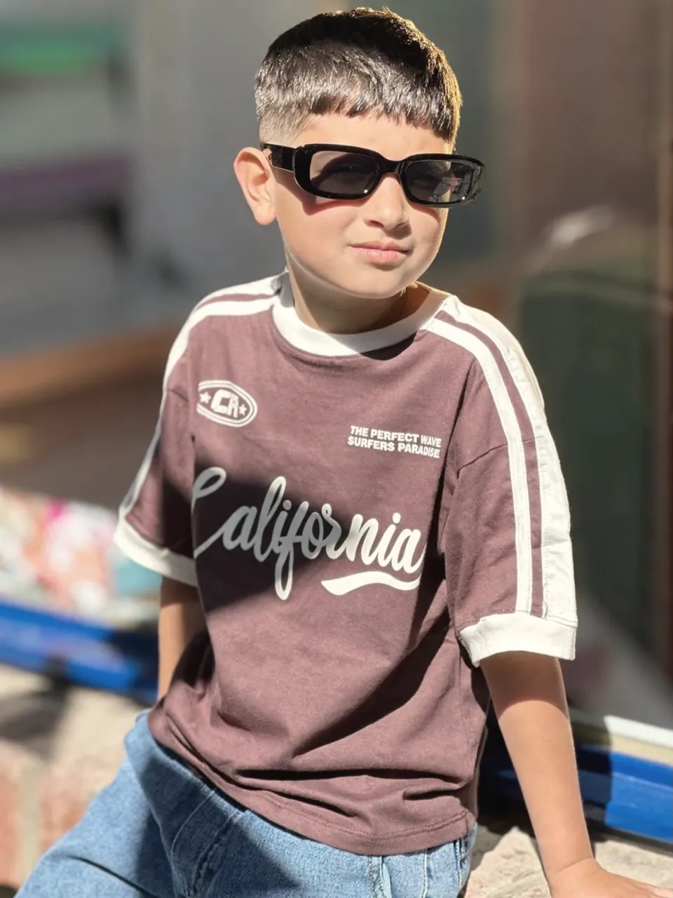 Chico con anteojos de sol y remera bordó California con vivos blancos, sentado al sol